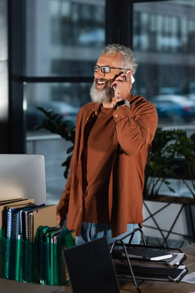 Alegre gerente de pelo gris hablando en el teléfono celular en la oficina por la noche - foto de stock