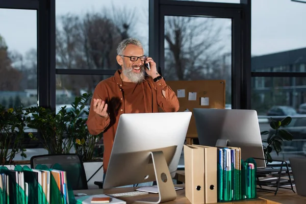 Positive grauhaarige Manager sprechen am Handy in der Nähe von Computern und Papierkram im Büro am Abend — Stockfoto