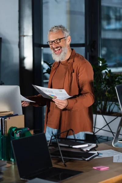 Cheerful businessman in eyeglasses holding papers with charts near computer in office in evening — Stock Photo