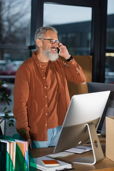 Grey haired businessman in casual clothes talking on cellphone near papers and commuter in office — Stock Photo
