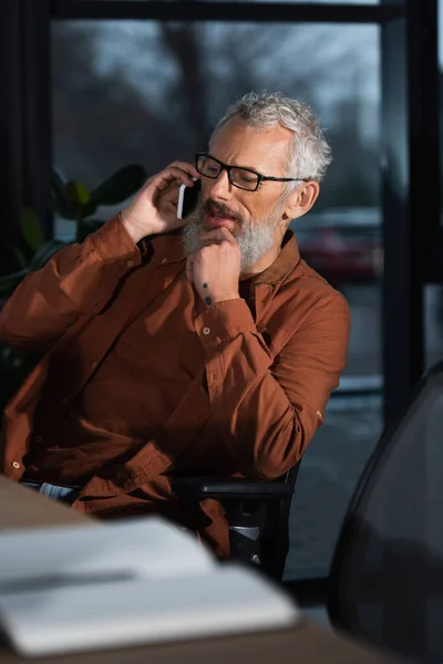 Mature businessman talking on smartphone near blurred notebook in office in evening — Stock Photo