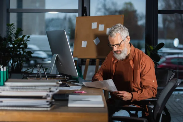Hombre de negocios de pelo gris que trabaja con papeles mientras está sentado en el lugar de trabajo cerca del monitor de computadora en la oficina - foto de stock