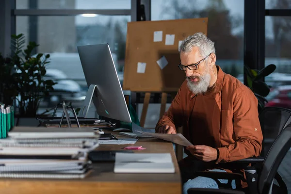 Homem de negócios barbudo em óculos que trabalham com documento perto de cadernos e monitor de computador no escritório à noite — Fotografia de Stock