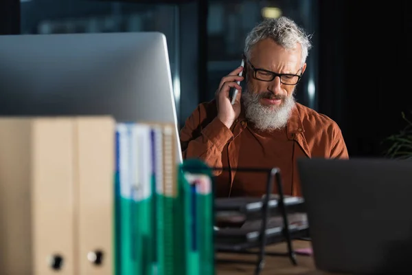 Grave homme d'affaires aux cheveux gris parler sur smartphone près des ordinateurs et des dossiers flous dans le bureau — Photo de stock