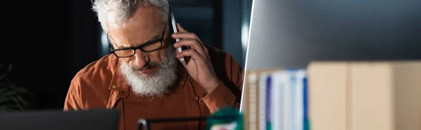 Bearded businessman in eyeglasses talking on smartphone near blurred computer monitor and folders, banner — Stock Photo
