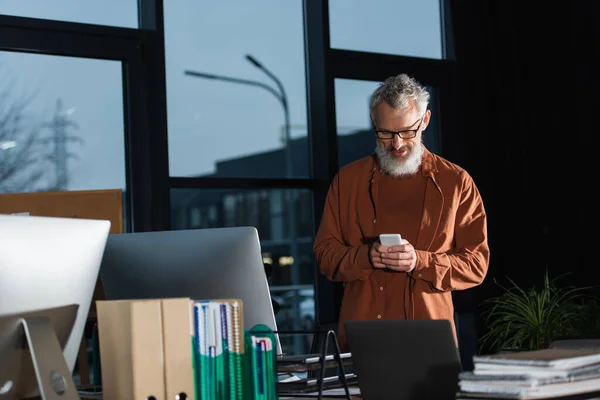 Grey haired businessman using smartphone while standing at workplace near computers in office — Stock Photo