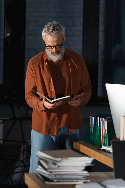 Middle aged businessman standing near documents and computer monitor and looking in notebook — Stock Photo