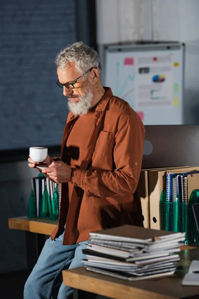 Бородатый бизнесмен среднего возраста с чашкой кофе и смартфоном, стоящий рядом с папками и ноутбуками в офисе — стоковое фото