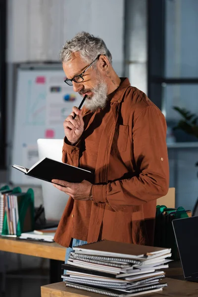 Thoughtful grey haired businessman in eyeglasses holding pen and looking in notebook in office — Stock Photo