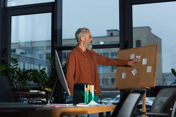 Вид збоку бізнесмена середнього віку, який дивиться на паперові нотатки на дошці під час роботи ввечері в офісі — стокове фото
