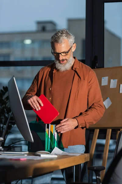 Middle aged manager in eyeglasses standing with notebook near computer monitor in evening in office — Stock Photo