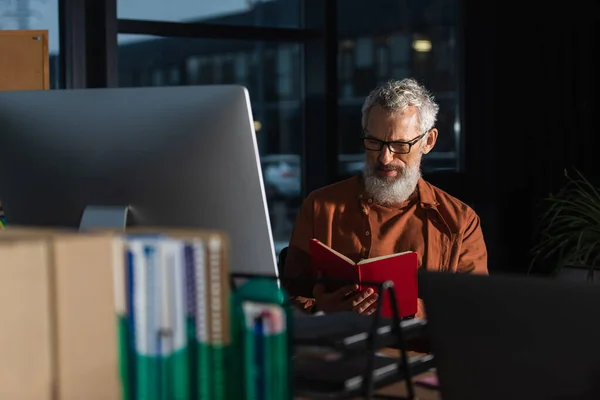 Mature businessman in eyeglasses looking in notebook near computer monitor in dark office — Stock Photo