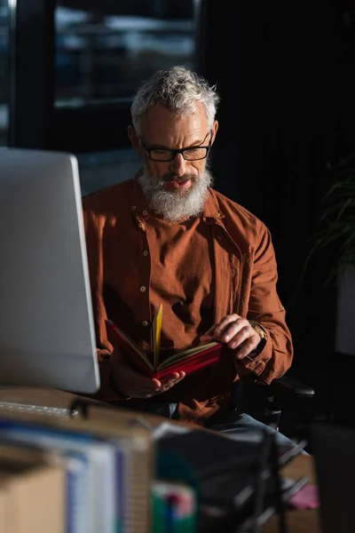 Thoughtful bearded businessman looking in notebook near computer monitor on blurred foreground — Stock Photo