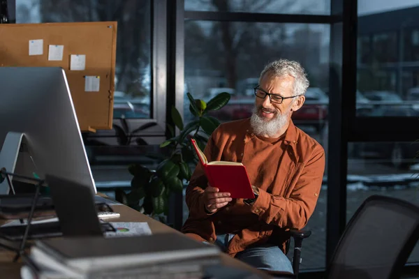 Feliz hombre de negocios de mediana edad sentado con copybook cerca del monitor de la computadora en el lugar de trabajo en la oficina - foto de stock