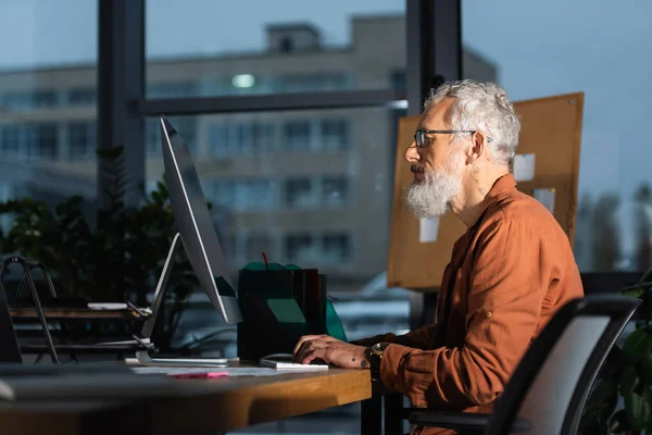 Боковой вид бородатого бизнесмена в очках, печатающего на ноутбуке в офисе вечером — стоковое фото