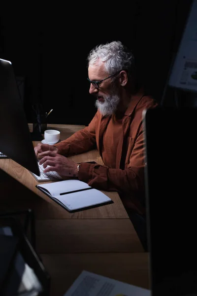 Bearded grey haired businessman typing on computer near notebook at night in office — Stock Photo