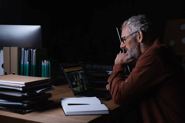 Side view of bearded man in eyeglasses working on laptop near pile of copybooks at night in office — Stock Photo