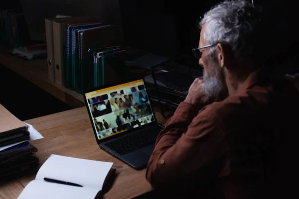 Middle aged businessman looking at laptop while sitting near blank notebook on office desk at night — Stock Photo