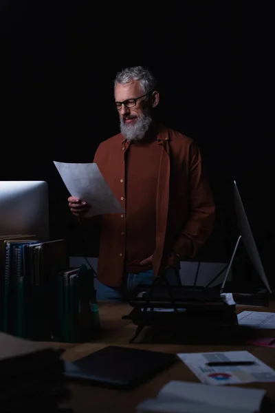 Bearded man standing with hand in pocket and looking at document near monitors in dark office — Stock Photo