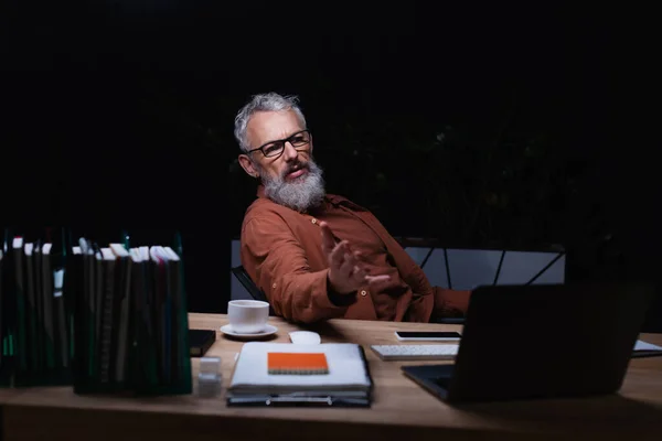 Bearded businessman in eyeglasses pointing with hand while talking during video chat on laptop in dark office — Stock Photo