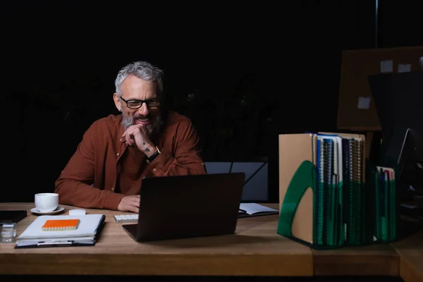 Allegro uomo d'affari dai capelli grigi che guarda il computer portatile mentre fa videochiamata di notte in ufficio — Foto stock