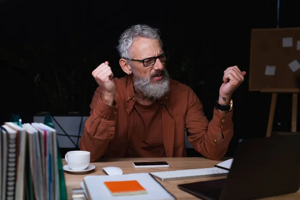 Раздраженный бизнесмен сжимает кулаки во время видеозвонка на ноутбук в офисе ночью — стоковое фото