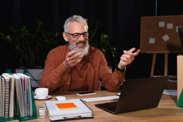 Allegro uomo d'affari barbuto che punta al computer portatile mentre ha videochiamata in ufficio di notte — Foto stock