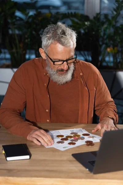 KYIV, UKRAINE - NOVEMBER 17, 2022: Bearded businessman in eyeglasses looking at bitcoins near notebook and laptop at workplace — Stock Photo