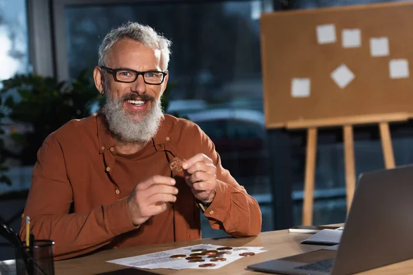 Bearded businessman in eyeglasses smiling at camera near bitcoins and laptop at workplace — Stock Photo