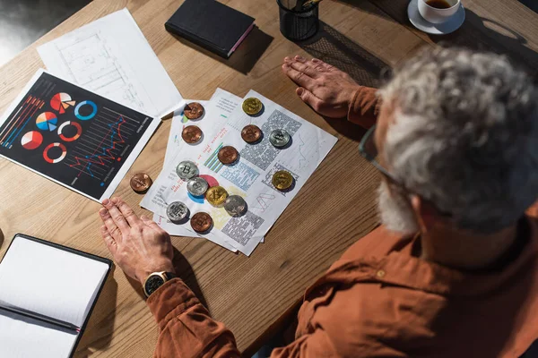 KYIV, UKRAINE - NOVEMBER 17, 2022: top view of mature businessman near bitcoins and documents with charts on wooden table — Stock Photo