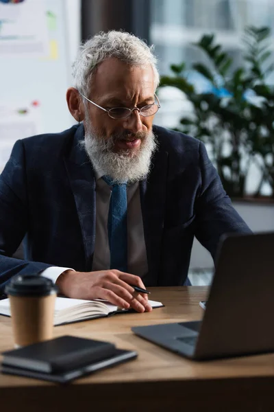Middle aged bearded man sitting with notebook and pen near laptop and blurred paper cup — Stock Photo