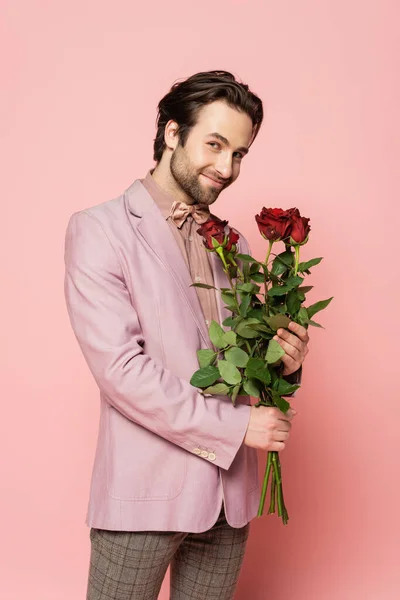 Anfitrión positivo de evento en chaqueta y pajarita sosteniendo rosas sobre fondo rosa - foto de stock