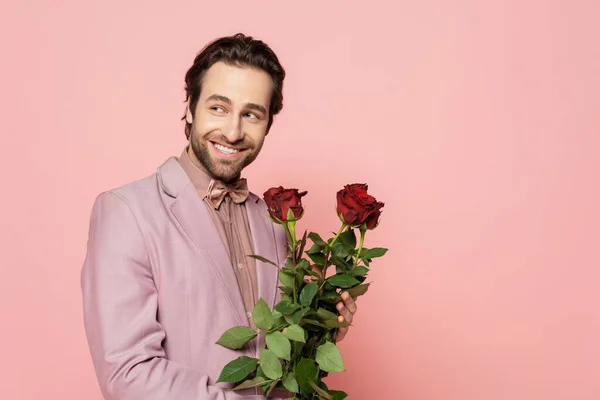 Alegre anfitrión del evento sosteniendo rosas rojas y mirando hacia otro lado aislado en rosa - foto de stock