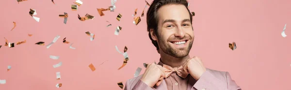 Positive host of event adjusting bow tie near confetti on pink background, banner — Stock Photo