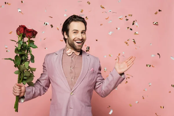 Excited host of event holding roses and pointing with hand under confetti on pink background — Stock Photo