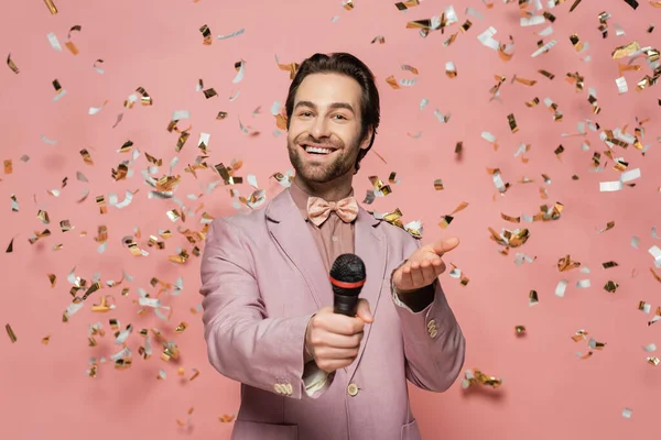 Smiling host of event holding microphone and pointing at camera under confetti on pink background — Stock Photo