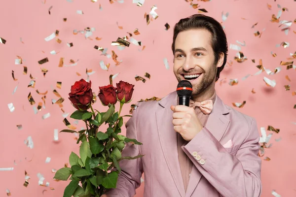 Cheerful host of event talking while holding microphone and roses on pink background with confetti — Stock Photo