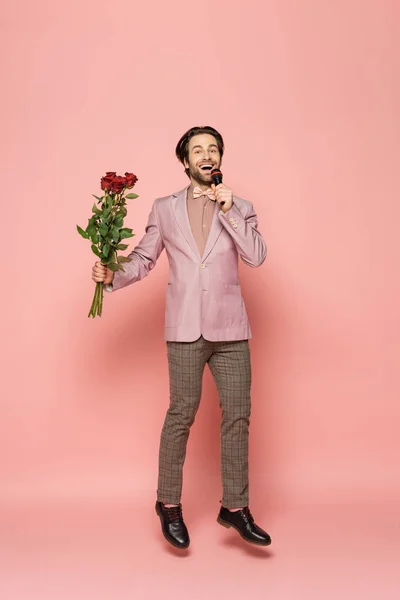 Positive host of event holding flowers and microphone while jumping on pink background — Stock Photo