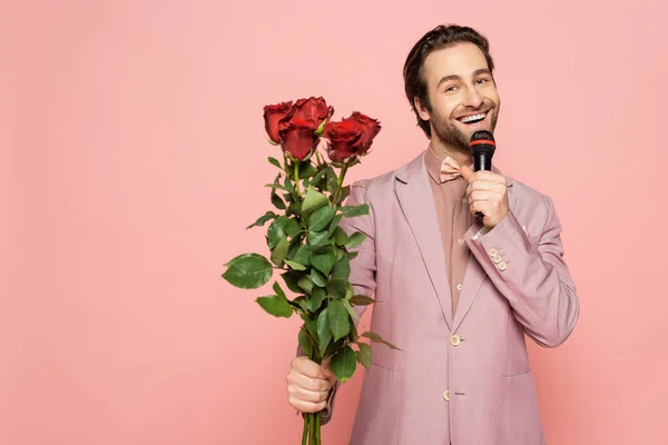 Cheerful host of event holding microphone and bouquet of roses isolated on pink — Stock Photo