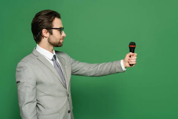 Side view of newscaster in suit holding microphone and looking away on green background — Stock Photo