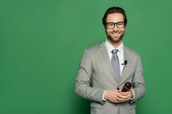 Fröhlicher Nachrichtensprecher in Anzug und Brille mit Mikrofon auf grünem Hintergrund — Stockfoto