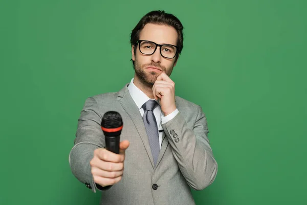 Pensive news anchor in suit and eyeglasses holding microphone isolated on green — Stock Photo