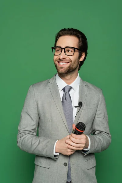 Happy news anchor in suit and eyeglasses holding microphone isolated on green — Stock Photo