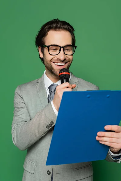 Щасливий інформаційний якір у костюмі та окулярах, що розмовляють мікрофоном, тримаючи буфер обміну ізольовано на зеленому — стокове фото