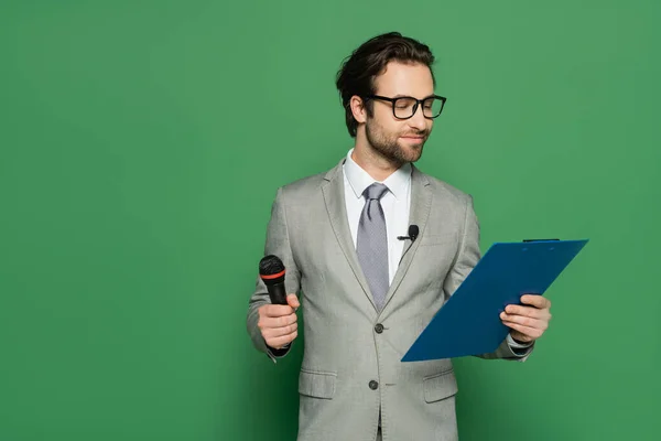 Broadcaster in suit and eyeglasses holding clipboard and microphone on green — Stock Photo