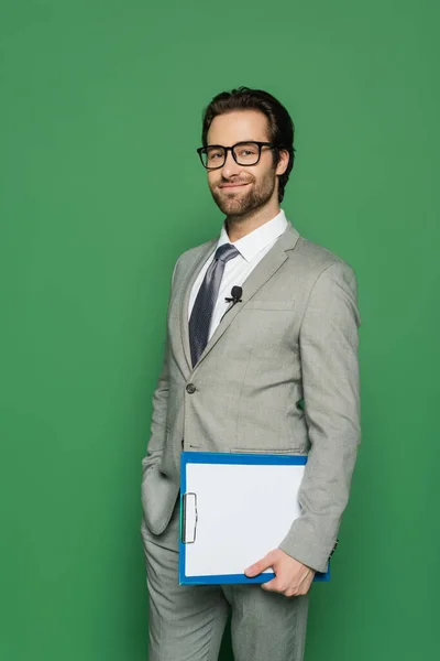 Happy news anchor in suit and eyeglasses holding clipboard while standing with hand in pocket isolated on green — Stock Photo