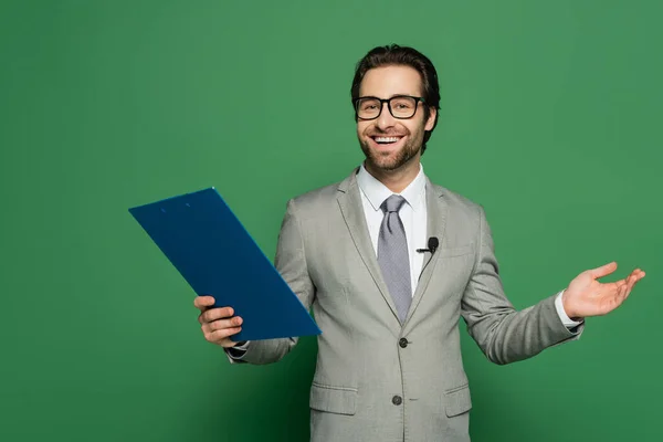Frohe Botschaft Anker in Anzug und Brille hält Klemmbrett, während gestikuliert auf grünem Hintergrund — Stockfoto