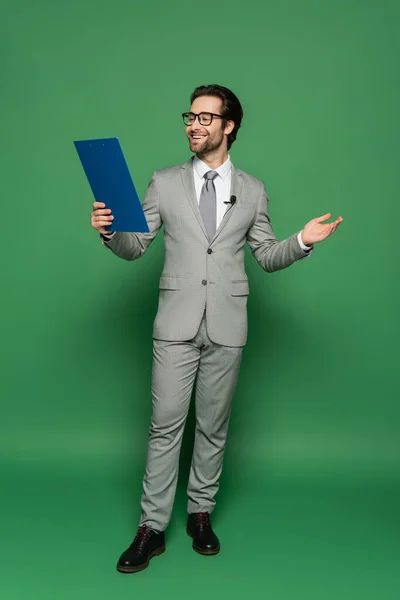 Full length of cheerful news anchor in suit and eyeglasses holding clipboard while gesturing on green — Stock Photo