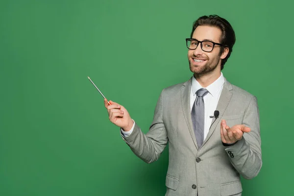 Alegre presentador de noticias en traje y gafas sonriendo mientras señala con lápiz sobre fondo verde - foto de stock