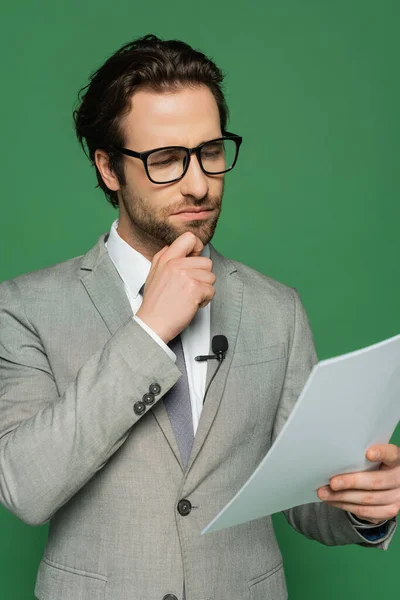 Reflexivo presentador de noticias en gafas y traje mirando papel en blanco aislado en verde - foto de stock